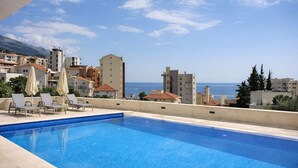 A swimming pool with lounge chairs and umbrellas, overlooking a coastal cityscape with buildings and a clear sky. - Residance with pool ,sauna and free garrage (Budva)