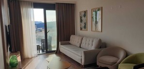 Ein Wohnzimmer mit einer Couch, einem Sessel, einem kleinen Tisch, einem Fernseher und einer Schiebetür zu einem Außenbereich.