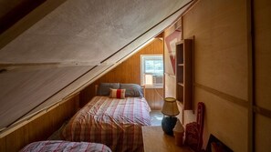 A small attic bedroom with a sloped ceiling, a window, a bed with a plaid bedspread, a bedside lamp, and a wall-mounted shelf. - Memory Island on Lamoka (Bradford)