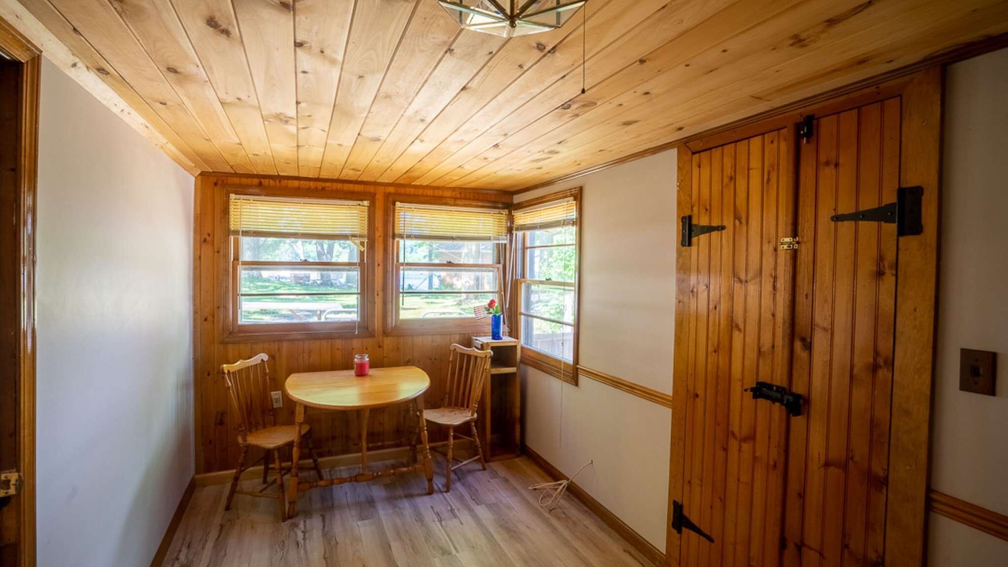 Una habitación con paneles de madera, una mesa redonda y dos sillas, una ventana con persianas y una puerta de madera.