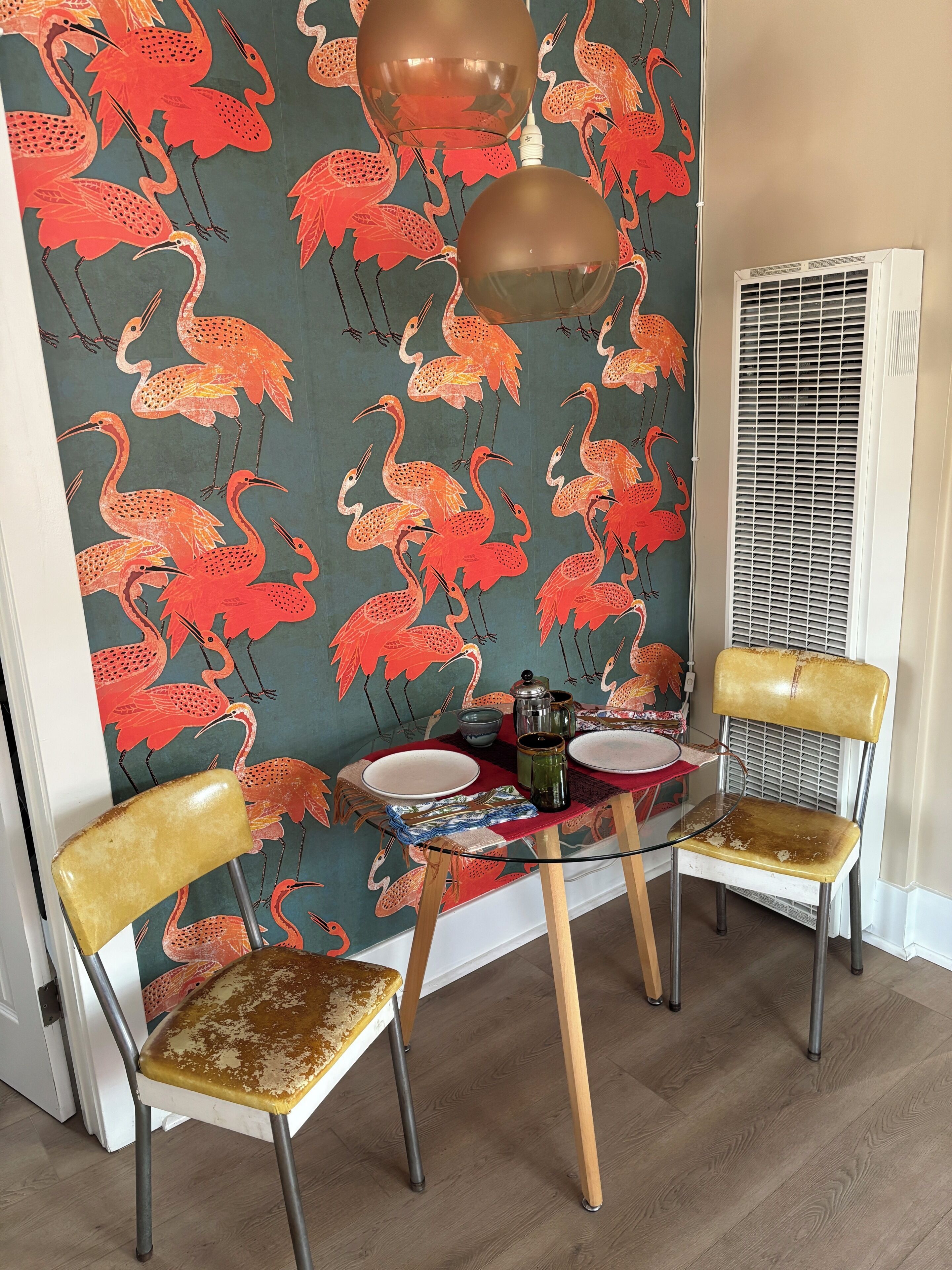 A dining area with a table set for two, two vintage-style chairs, a decorative wall with a flamingo pattern, and a ceiling light fixture.