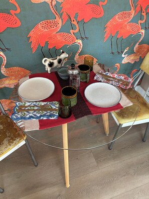 Uma mesa de jantar posta com pratos, copos e um jarro de leite, rodeada por cadeiras com almofadas estampadas.
