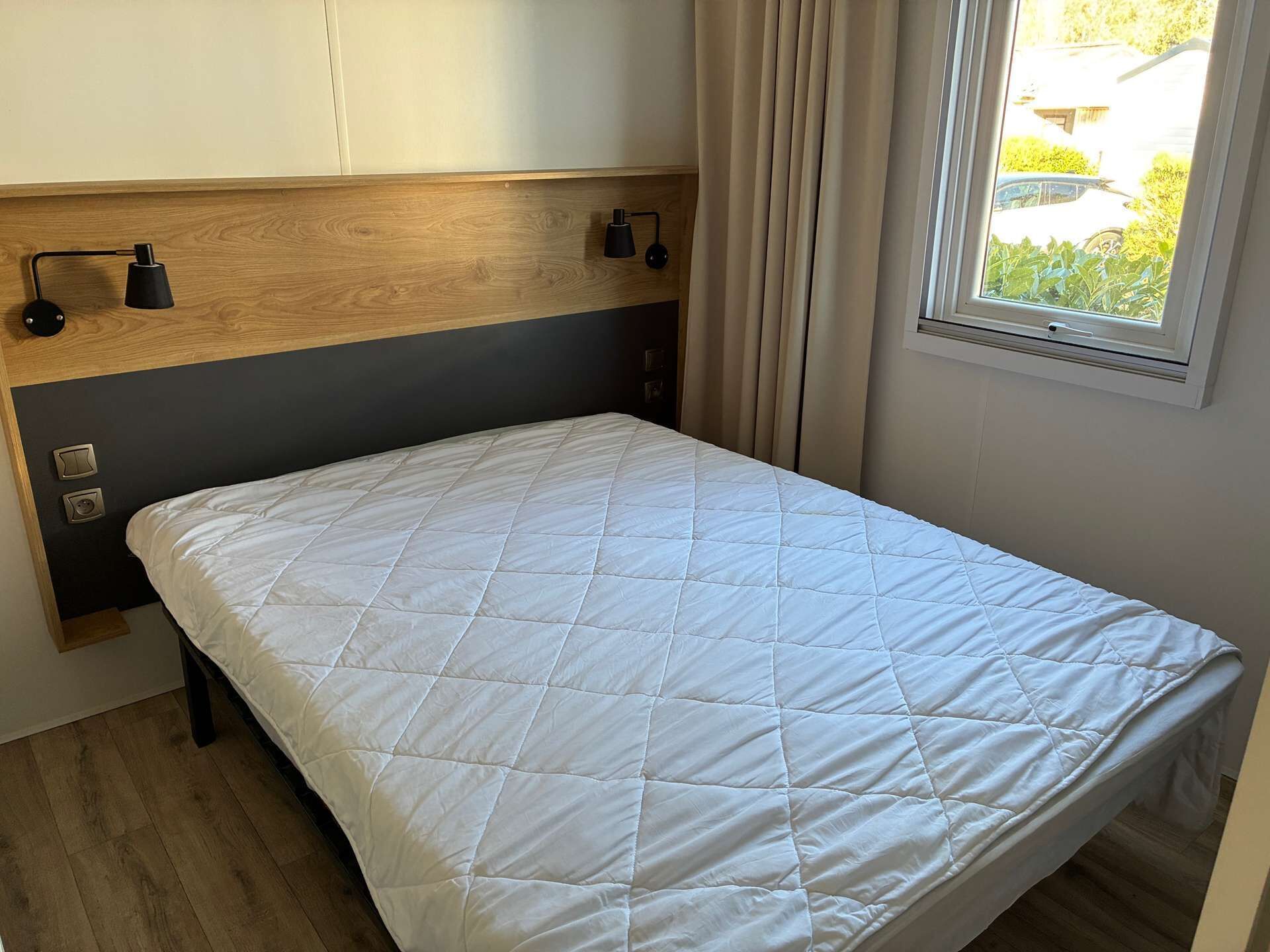 A single bed with a quilted white cover, a wooden headboard, and a window with curtains.