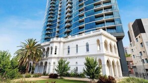 Un immeuble de grande hauteur doté d’une façade moderne en verre, d’une base de style classique avec des fenêtres en arc de cercle et d’un jardin bien entretenu.