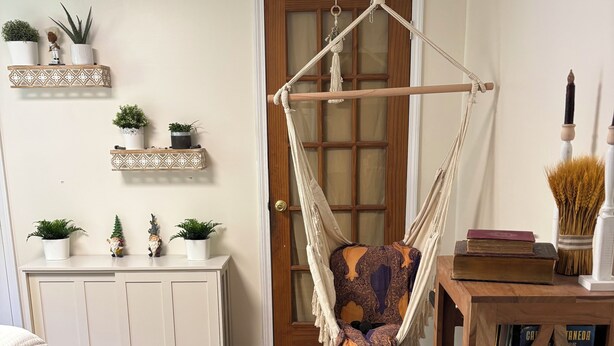 Una habitació amb sòl de fusta, una cadira penjant, un moble blanc amb plantes i una taula auxiliar de fusta amb llibres.