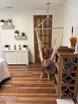 A room with a wooden floor, a hanging chair, a white cabinet with plants, and a wooden side table with books.