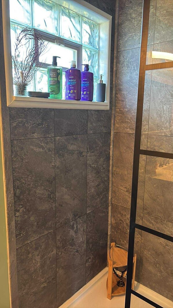 A bathroom with dark tiles, a window with toiletries, and a wooden stool.