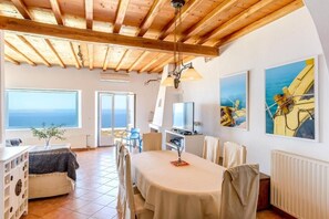 Ein geräumiges Zimmer mit Holzbalkendecke, einem Esstisch, Stühlen, einem Fernseher und Kunstwerken an den Wänden.