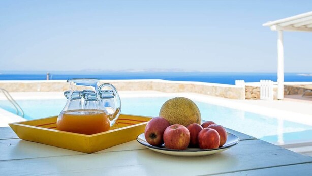 A table with a pitcher of orange juice, a plate with apples and a melon, and a pool with a view of the sea. - Lighthouse Villa w Private Pool & 220° Sea View (Faros Armenistis)