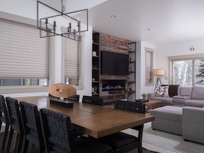 A modern living room with a fireplace, a dining area with a wooden table and chairs, a sofa, and a television mounted on a stone wall.