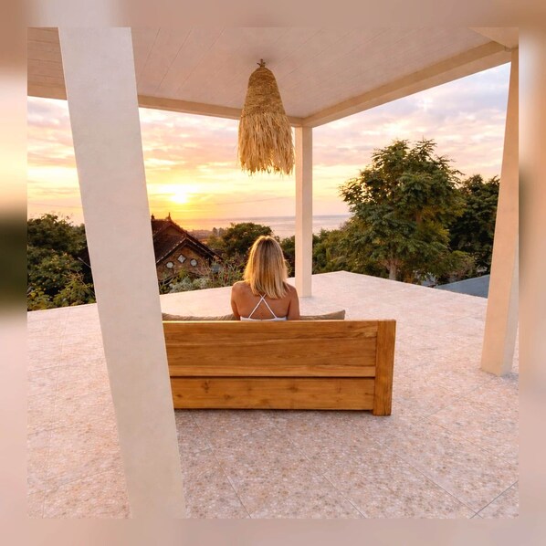 Una mujer sentada en un banco de madera, con vista a un atardecer y árboles.
