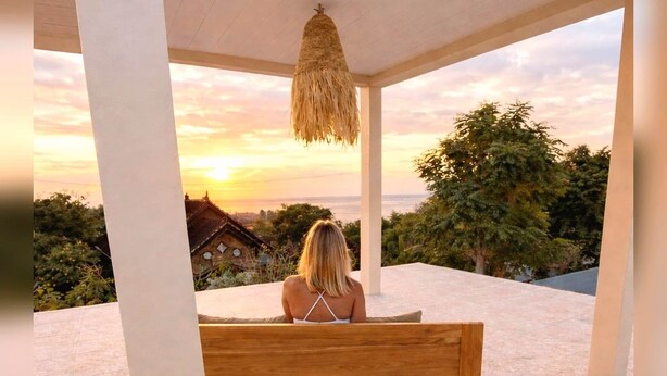A woman sitting on a wooden bench with a view of a sunset and trees. - Thula Lembongan Villa 3 (Lembongan)