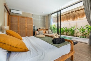 A modern bedroom with a large bed, a small kitchenette, and a view of a pool and greenery. - Thula Lembongan Villa 3 (Lembongan)
