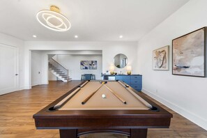 Una mesa de billar en una habitación con escalera, obras de arte en las paredes y una lámpara de techo moderna.