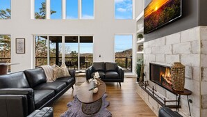 A modern living room with a fireplace, leather sofas, a coffee table, and large windows offering a view of the outdoors. - 🌄 Blue Ridge Mountain Escape | Scenic Mountain Getaway, Hot Tub & Game Room (Blue Ridge)
