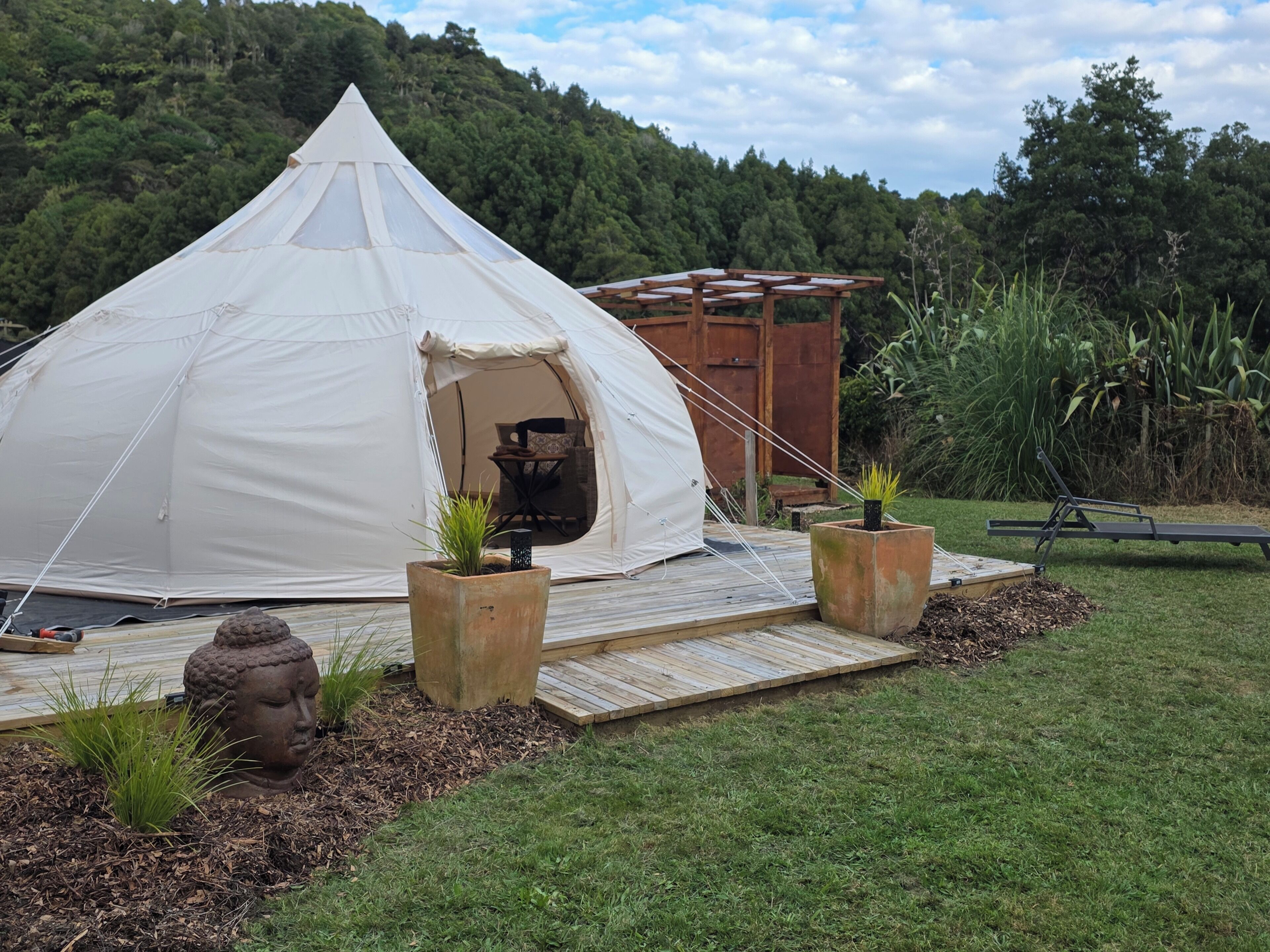 Una gran tenda blanca amb un terraç de fusta al davant, envoltada de vegetació i una estructura de fusta al fons.