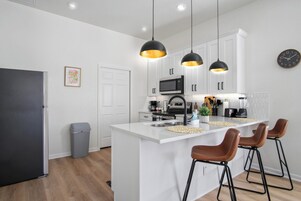 A modern kitchen with white cabinetry, a central island with stools, pendant lighting, a microwave, and a refrigerator.