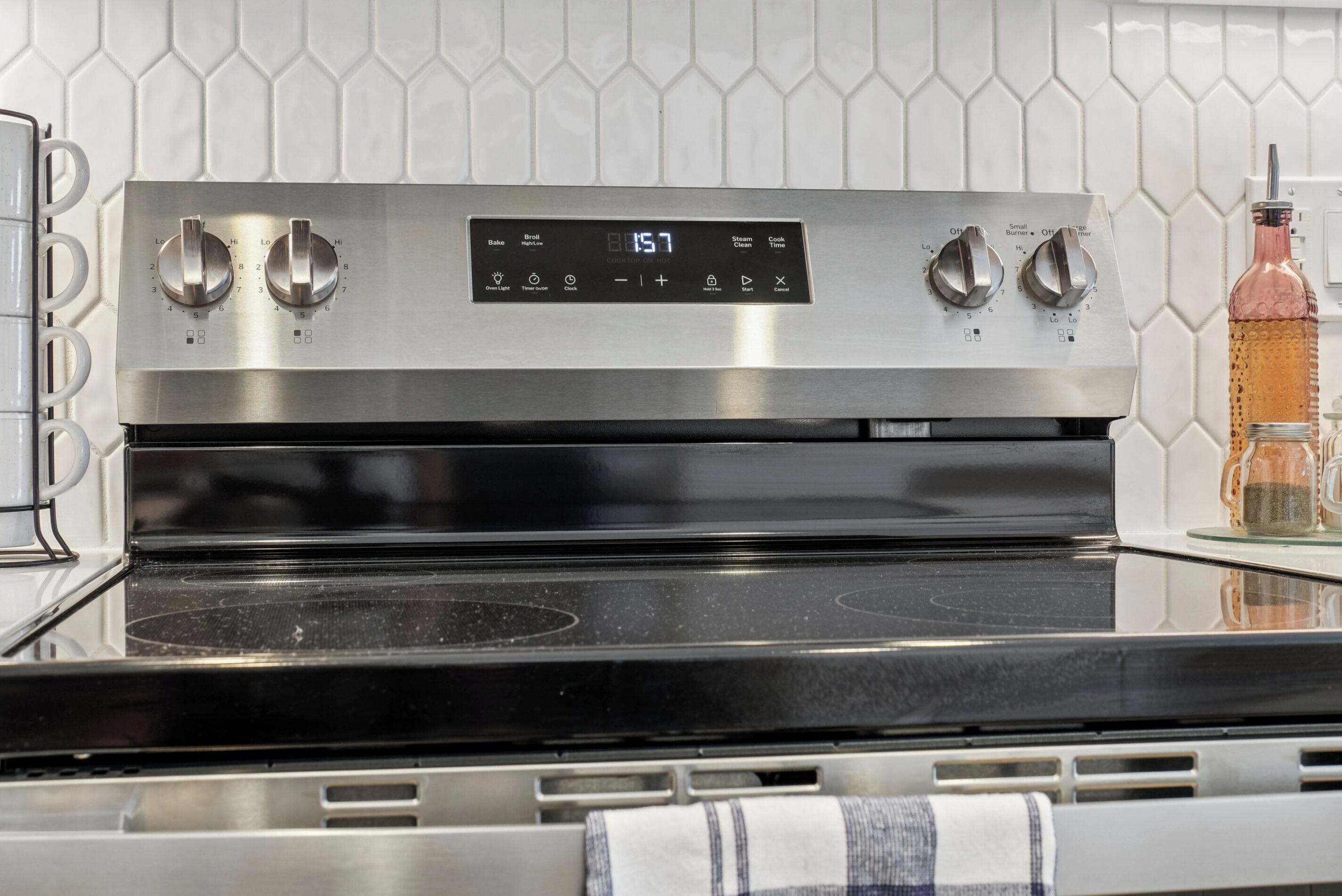 A stainless steel oven with a digital display, knobs, and a glass bottle on the counter.