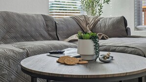 Un salon moderne avec un canapé gris à motifs matelassés, une table basse ronde sur laquelle sont posés des biscuits et une petite plante, ainsi qu’une fenêtre avec des stores.