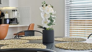 Une table à manger avec des sets de table en fibres tressées, un vase noir contenant des fleurs blanches et, au fond, un coin cuisine.