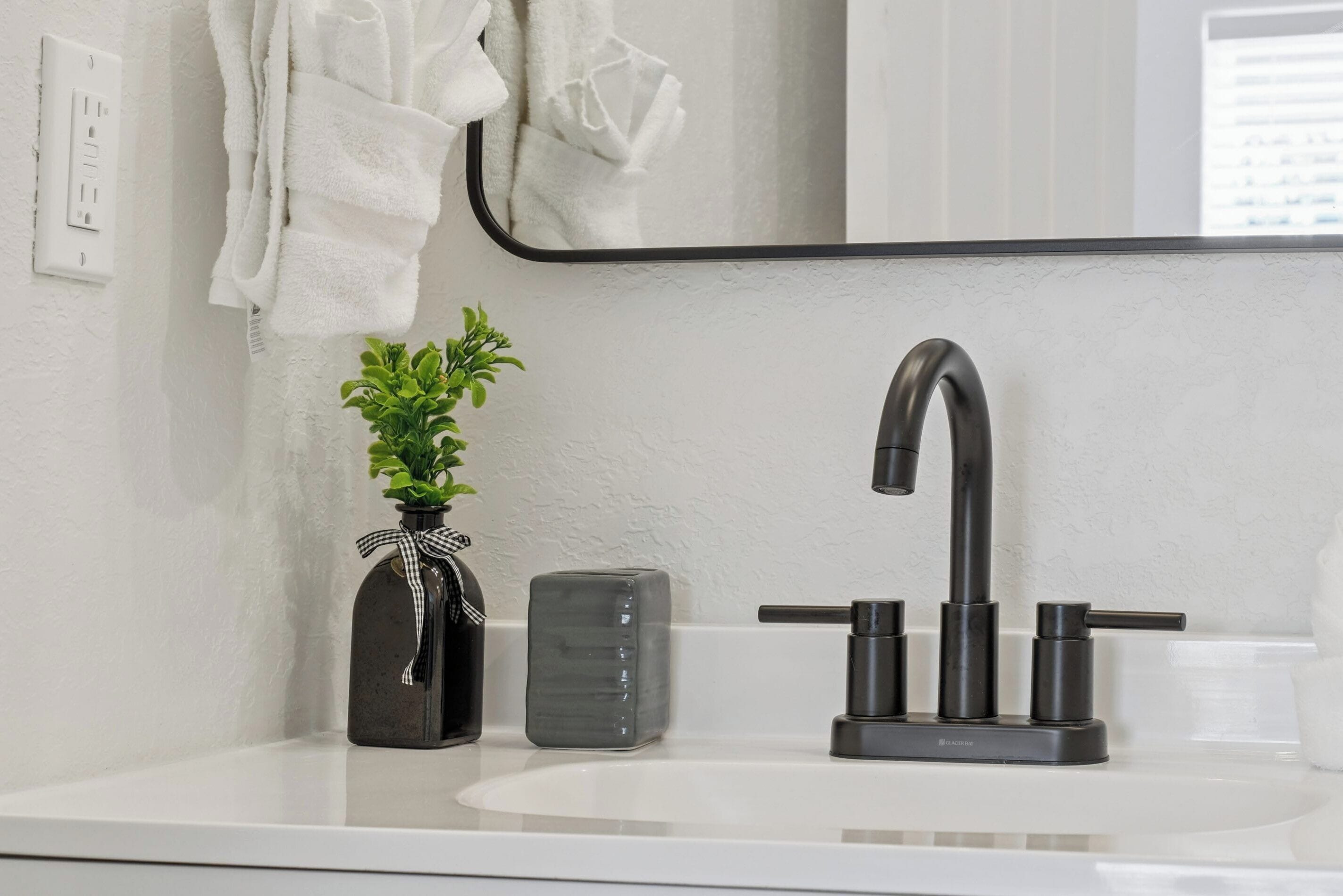 Un baño con un lavabo blanco, un grifo negro, una planta pequeña en maceta, un florero negro y una toalla blanca colgada en la pared.