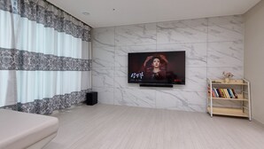 A modern living room with a flat-screen TV mounted on a marble wall, a beige sofa, a bookshelf, and patterned curtains. - Spacious 3BR Brand New Cozy Apt at Dongdaegu KTX Station (Daegu)