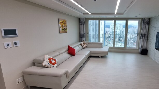 A modern living room with a sofa, a flat-screen TV, and a city view. - Spacious 3BR Brand New Cozy Apt at Dongdaegu KTX Station (Daegu)
