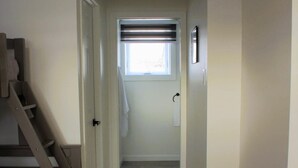 Un pasillo estrecho con una escalera de madera a la izquierda, una puerta con una toalla colgada y un ventanal con persianas al final.
