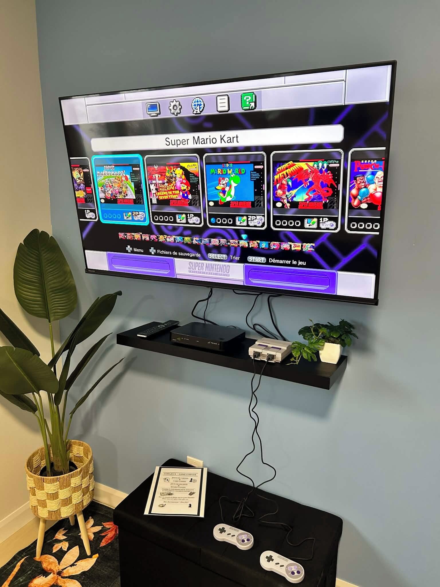 A television mounted on a wall displaying a Super Mario Kart game. Below the TV, there is a shelf with a remote control, a game console, and a small plant.