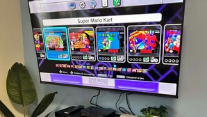 A television mounted on a wall displaying a Super Mario Kart game. Below the TV, there is a shelf with a remote control, a game console, and a small plant.