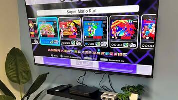 A television mounted on a wall displaying a Super Mario Kart game. Below the TV, there is a shelf with a remote control, a game console, and a small plant.