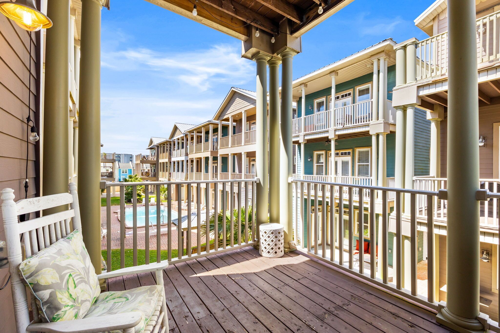 A wooden deck with a bench and a trash can, overlooking a pool and multi-story buildings with balconies.