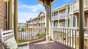 Une terrasse en bois avec un banc et une poubelle, donnant sur une piscine et des immeubles à plusieurs étages dotés de balcons.