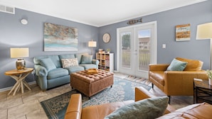 Una sala de estar con un sofá azul, un otoman marrón, un sillón de cuero color beige y una mesita de madera.