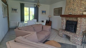 Een woonkamer met een bank, een open haard, een eettafel met stoelen, en een raam met gordijnen.