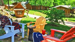 Ein rustikaler Sitzbereich im Freien mit Adirondack-Stühlen und einem kleinen Tisch, umgeben von Holzhütten und üppiger Vegetation.