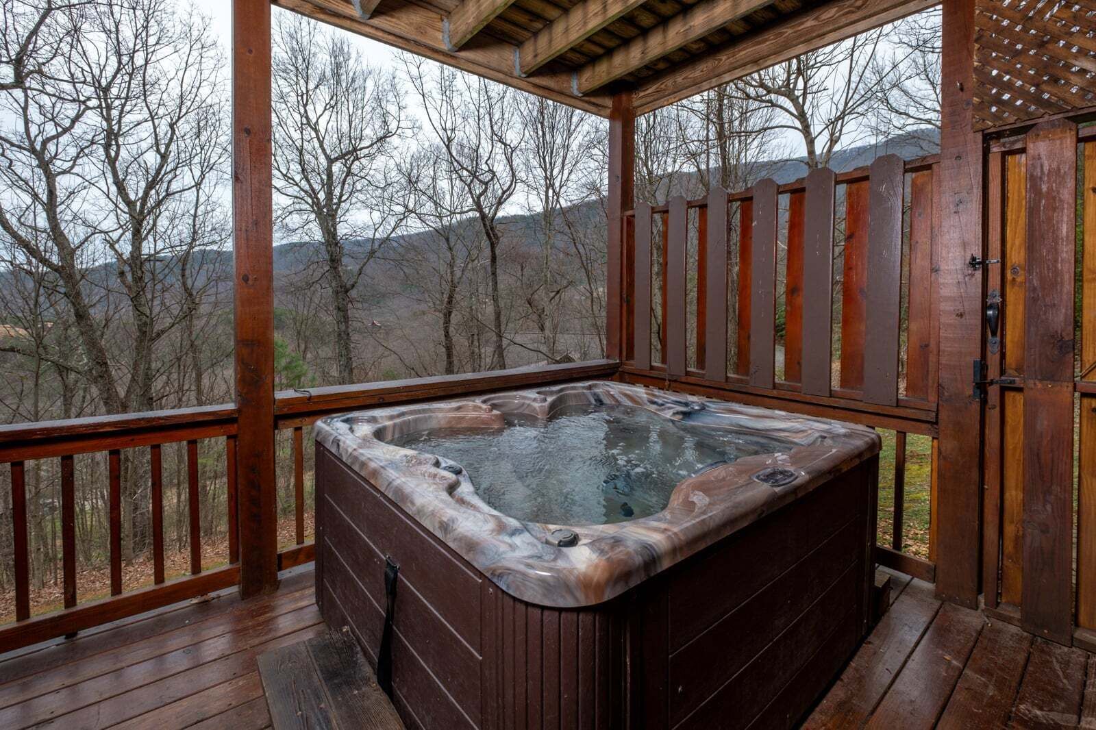 Un bain à remous sur une terrasse en bois, entouré d’une clôture en bois et doté d’une porte, le tout niché au milieu des arbres et des collines.