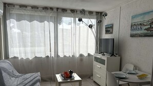 A room with a bed, a small table with a red mug, a TV on a stand, a painting on the wall, and a chair. - Air-conditioned studio by the beach - 3 guests (Port Camargue)