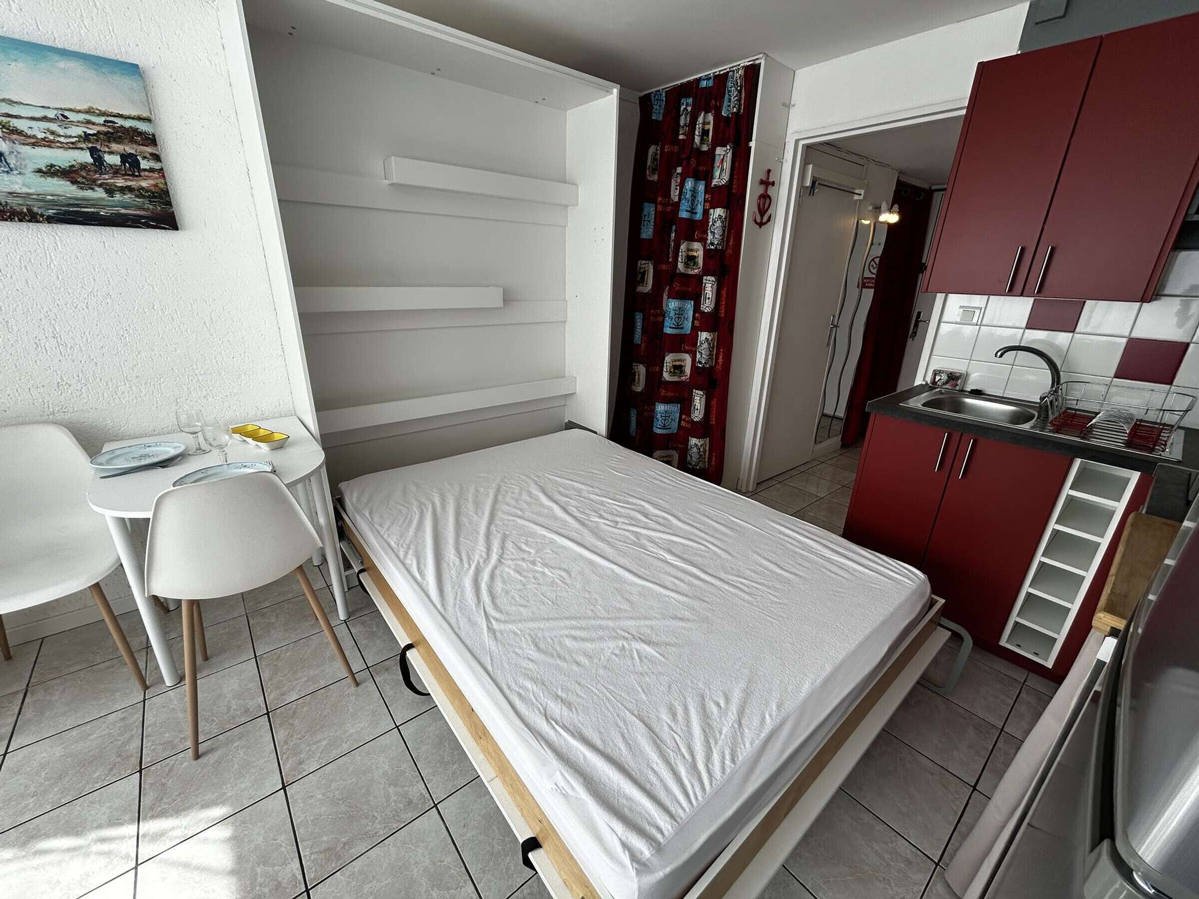 Une pièce de taille modeste comprenant un lit, une petite table à manger avec des chaises, un coin cuisine et un tableau accroché au mur.
