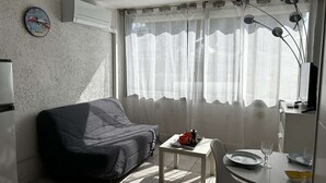 A small, well-lit room with a sofa, a table with a tray of items, a clock on the wall, and a window with sheer curtains. - Air-conditioned studio by the beach - 3 guests (Port Camargue)