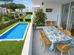 A poolside area with a rectangular pool, wicker chairs, a table with a patterned tablecloth, and a view of the surrounding greenery and buildings. - Lodging in La Fosca - Palamós (Palamós)