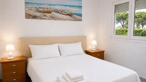 Ein Hotelzimmer mit einem Bett, zwei Nachttischen, einem Bild von Booten am Strand und einem Fenster mit Blick auf Bäume.