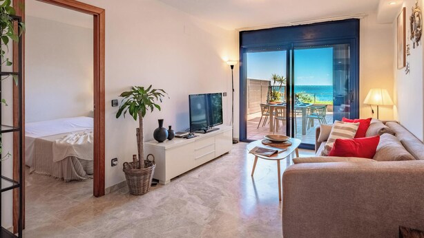 Una sala de estar moderna con televisor, sofá, mesa de comedor y vista al mar a través de un gran ventanal corredero.