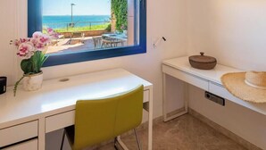 Una habitación con escritorio, una silla, ventana con vista al mar, un estante pequeño con un sombrero y un jarrón con flores.