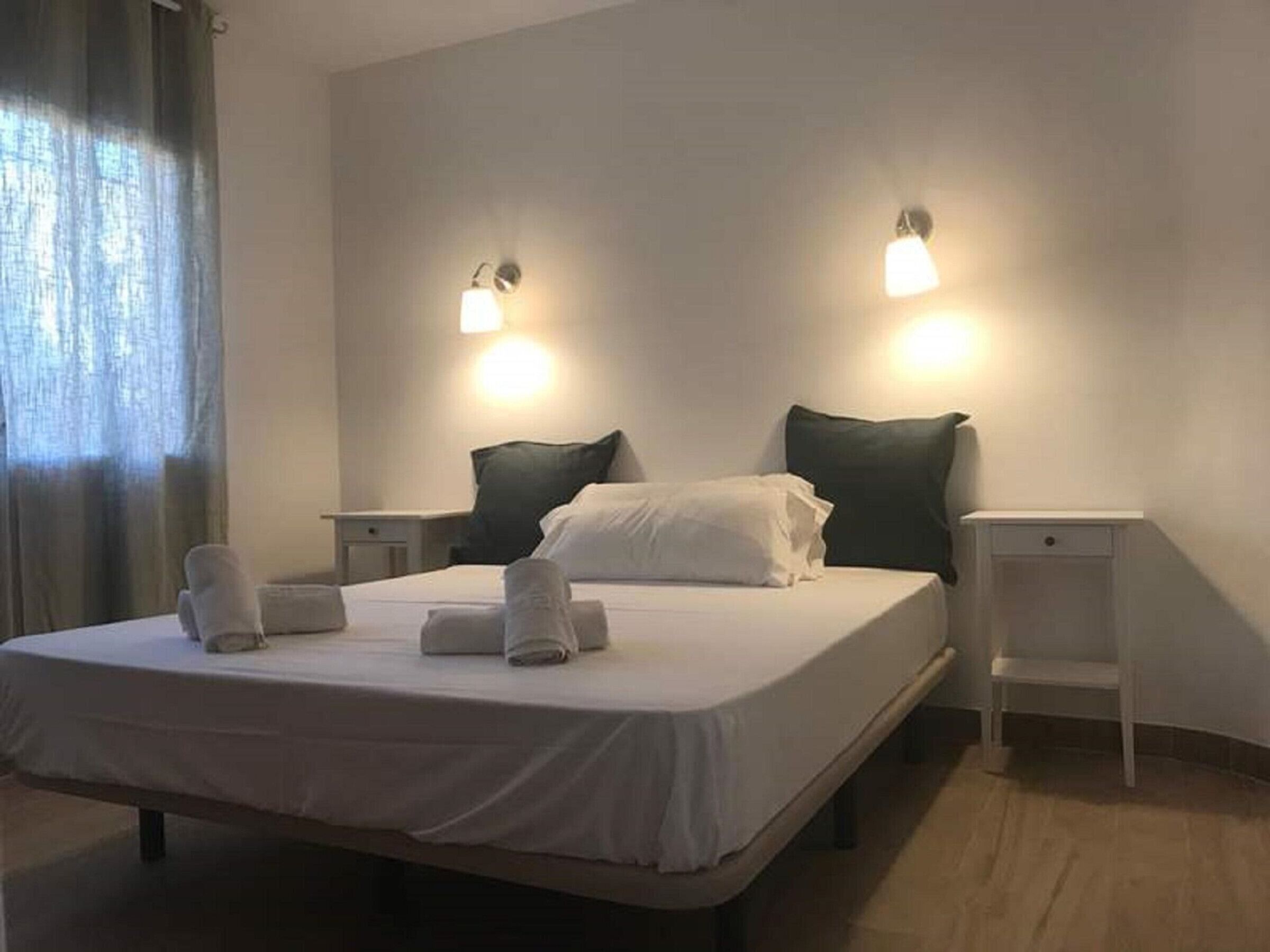 Ett hotellrum med en säng, två sängbord, ett litet bord, en lampa och ett fönster med gardiner.
