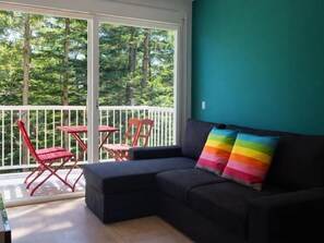 A room with a dark-colored sofa, a colorful pillow, a white railing, and a view of trees through large windows. - Quiet apartment near beach - pers. (Palamós)