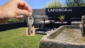 Una mano sosteniendo llaves frente a un letrero que dice 'LAFOSCA.es', con mobiliario exterior y una fuente.