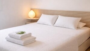 Una cama bien tendida con sábanas y almohadas blancas, una lámpara de noche y una pila de toallas dobladas.