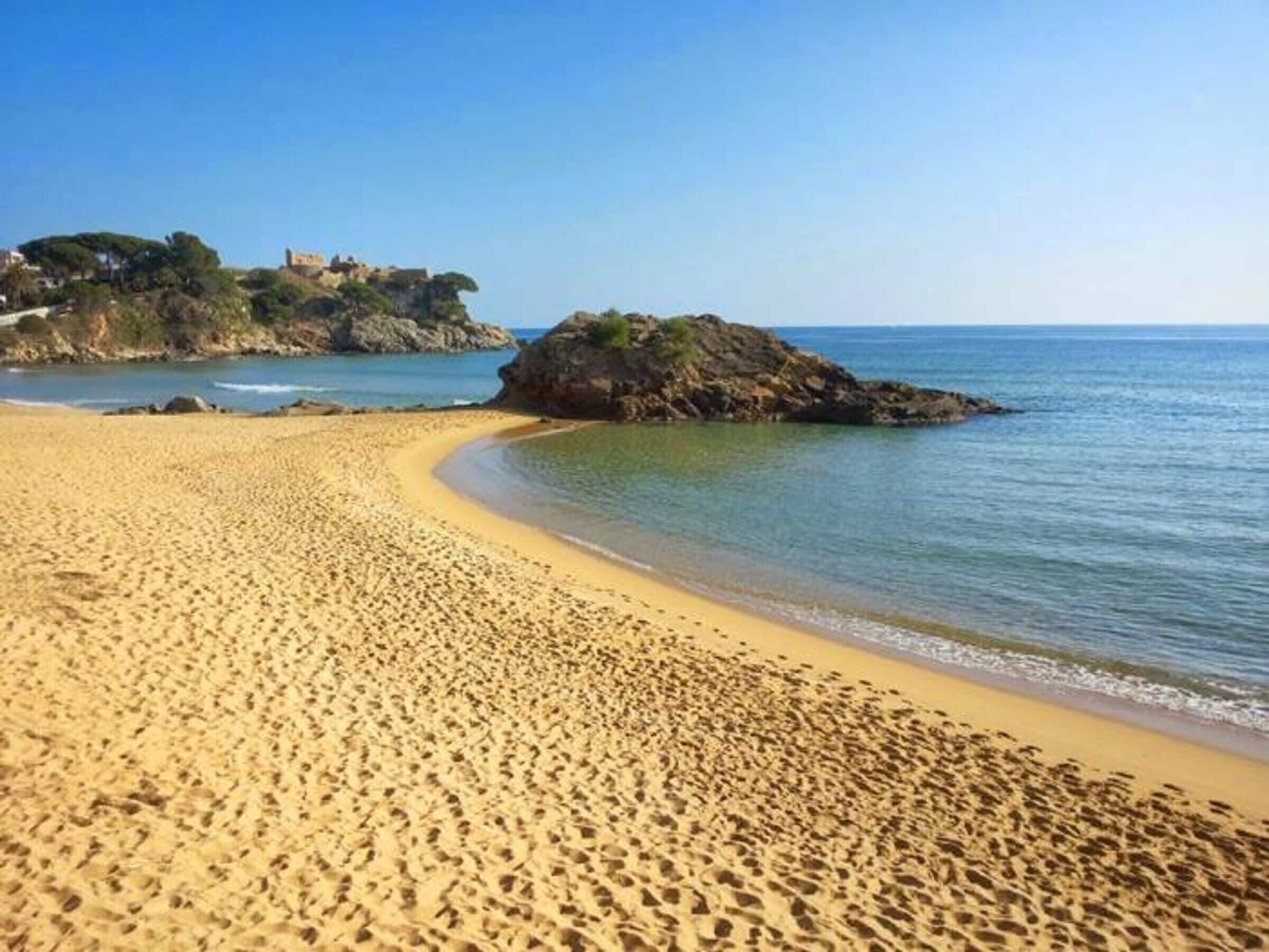 Ein sandiger Strand mit klarem, blauem Wasser, einem Felsen und einem Gebäude auf einem Hügel im Hintergrund.