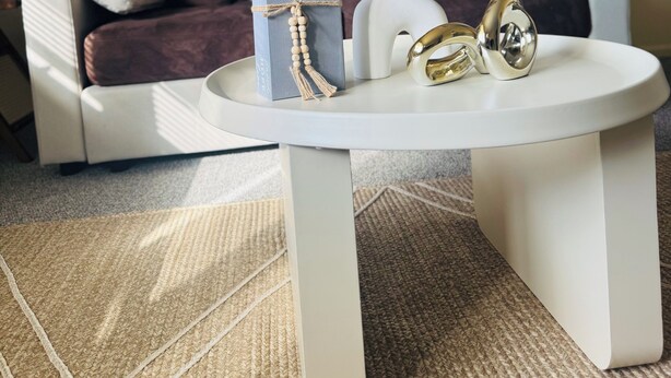 Una sala de estar con un sofá blanco, una mesa de centro redonda, almohadones decorativos, obras de arte en la pared y una alfombra tejida.